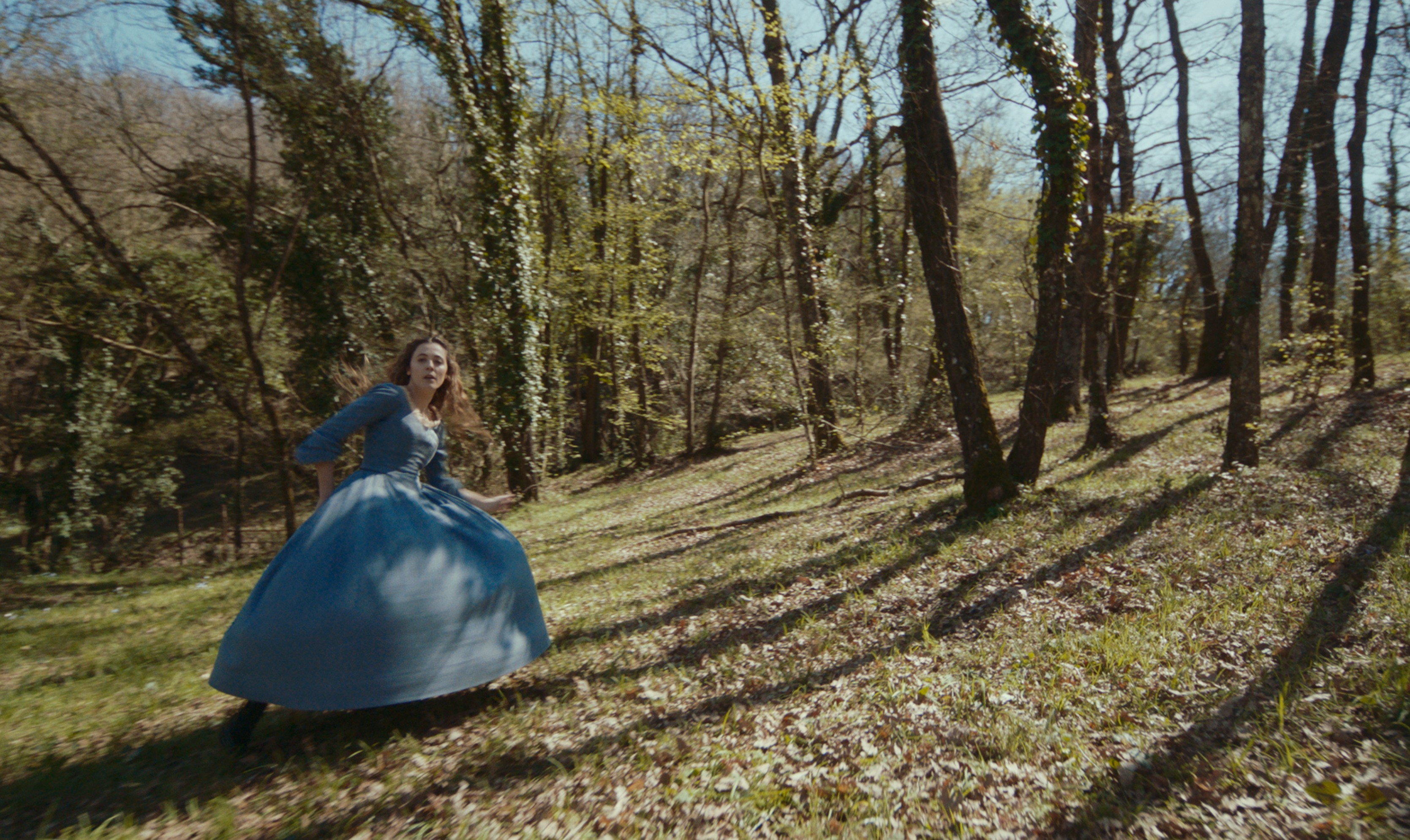 A woman in a blue dress turning in a sunlit woodland