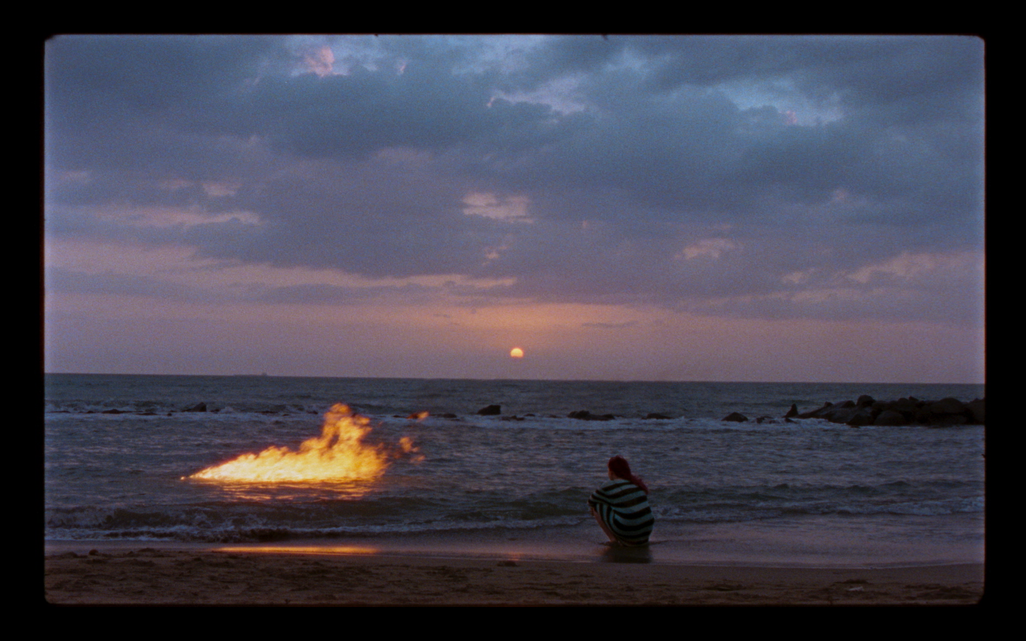 A figure beside the sea with fire on the water at sunset