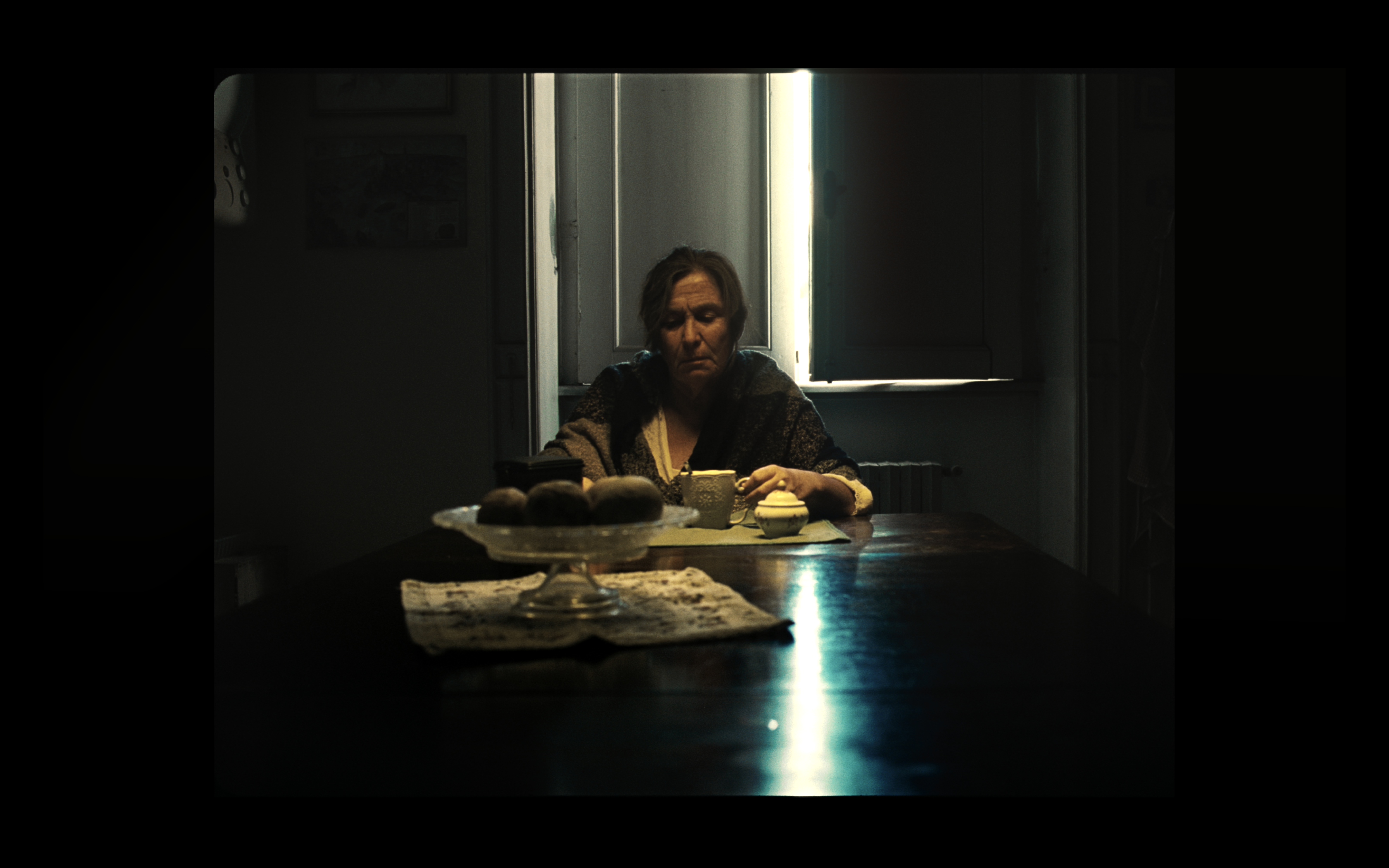 An older woman sitting at a table beside a shuttered window