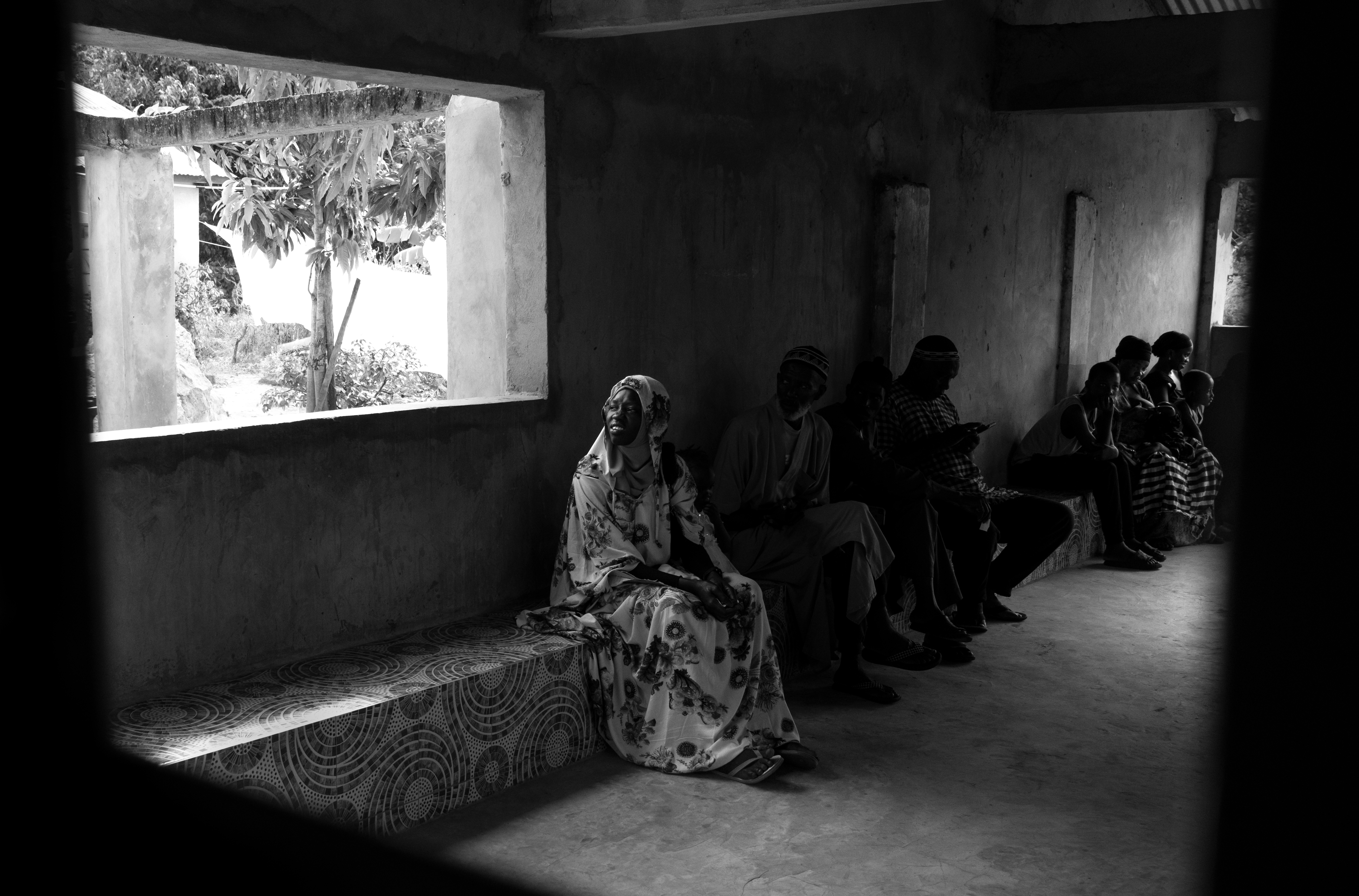 People seated along a wall beside an open window in black and white