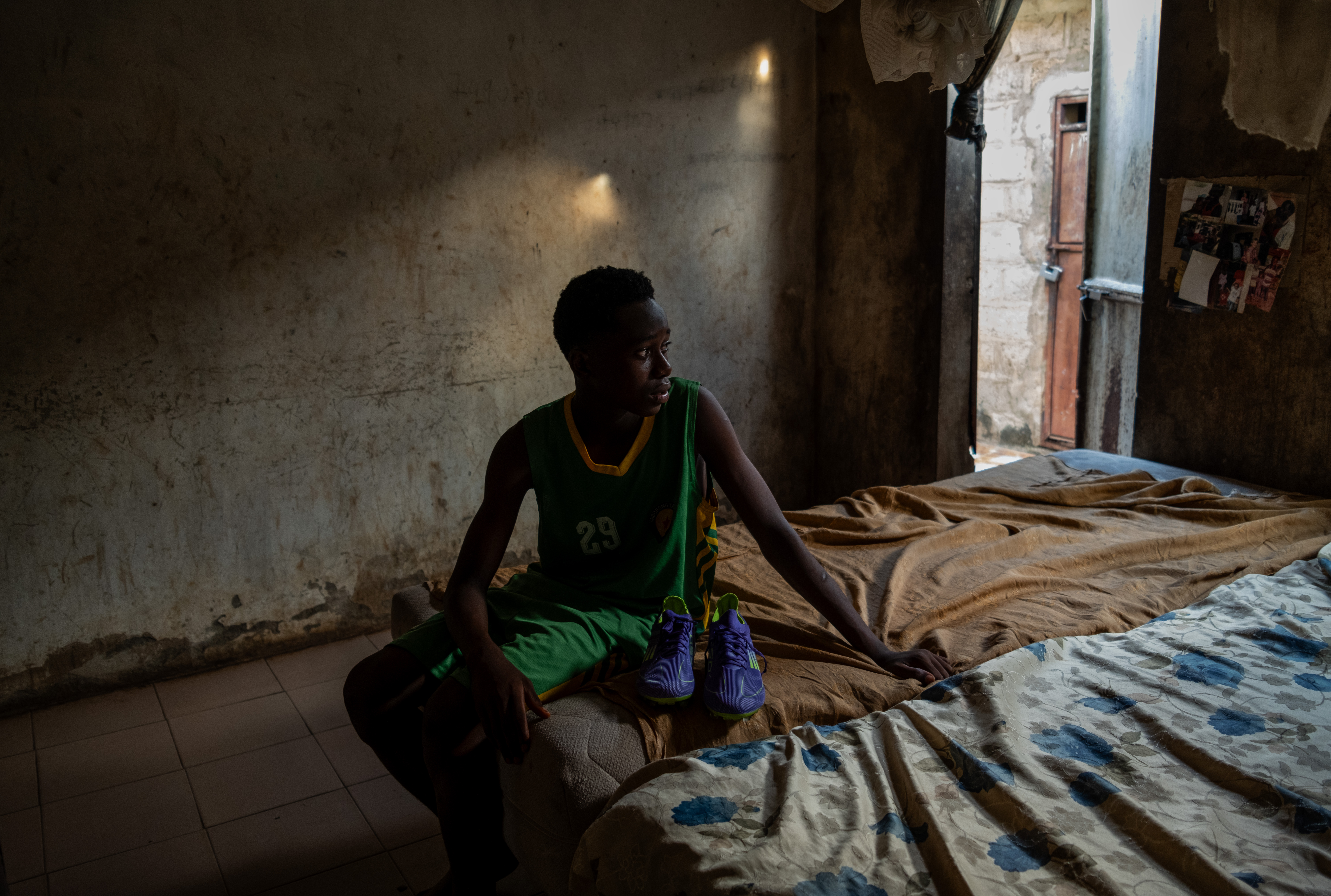A young person sitting on a bed beside a sunlit doorway