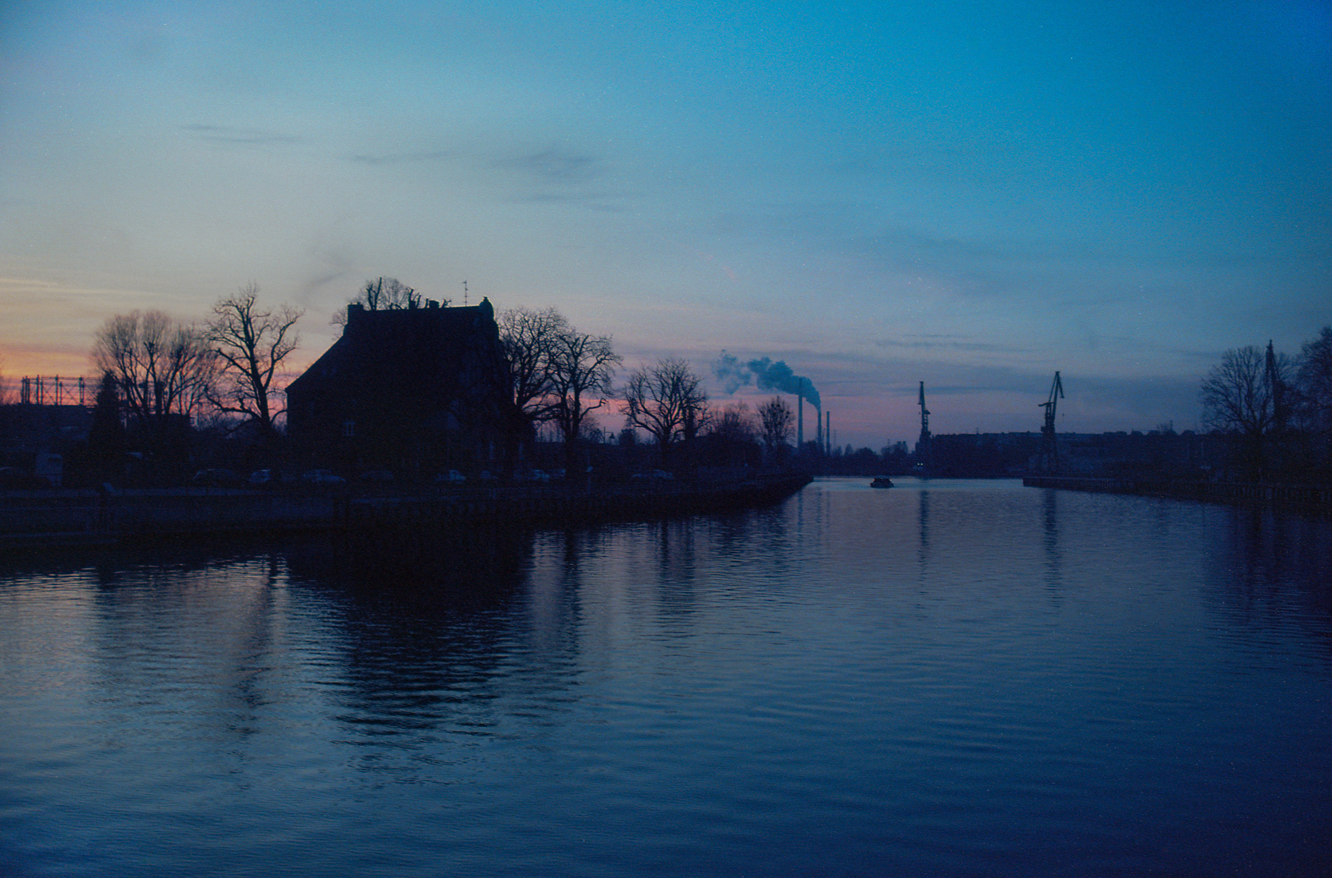 A quiet river and industrial silhouettes beneath a blue evening sky