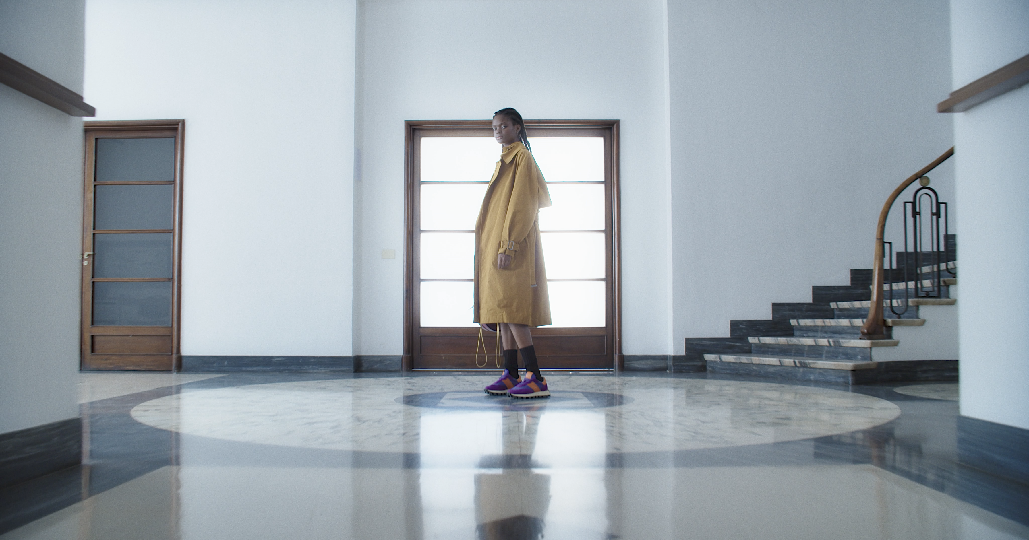 A woman wearing a yellow coat and sneakers in a bright hall