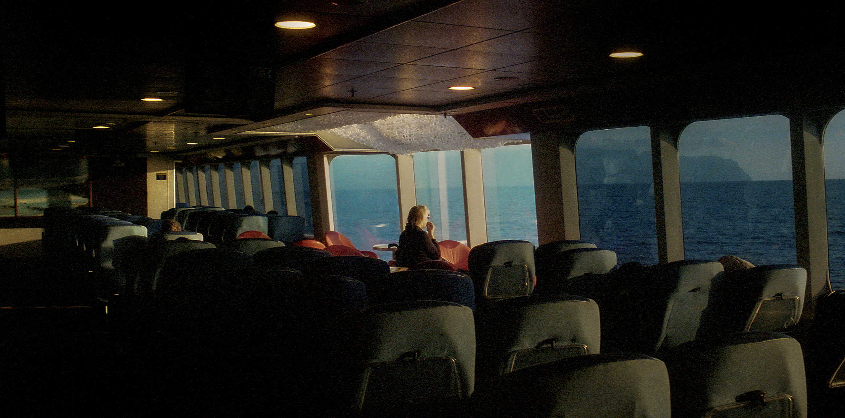 A passenger in warm sunlight beside the sea-facing windows of a ferry